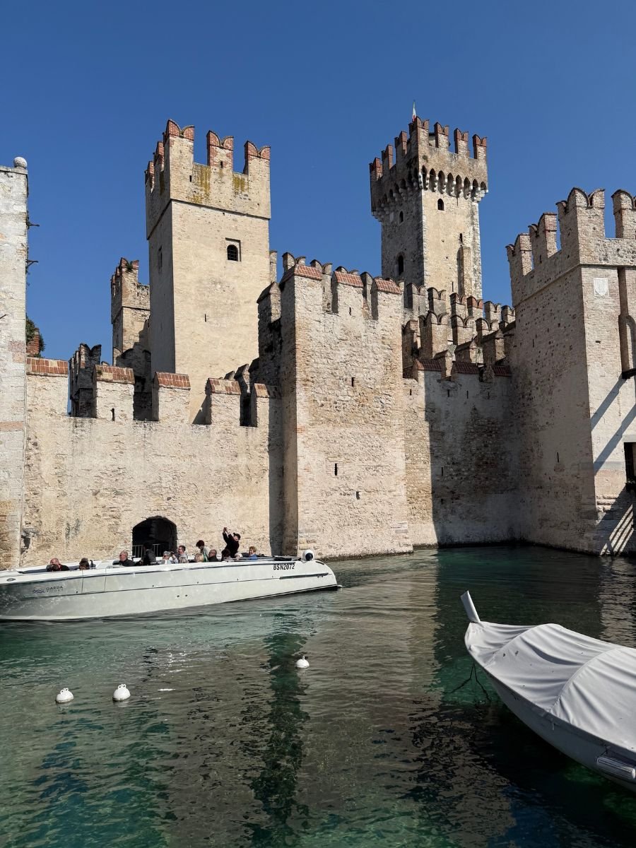 Scaliger Castle in Sirmione, Italy, with its medieval stone walls and crenellated towers rising above a clear blue moat. A white motorboat carrying tourists glides near the castle entrance, while another boat is docked nearby under a bright, cloudless sky.