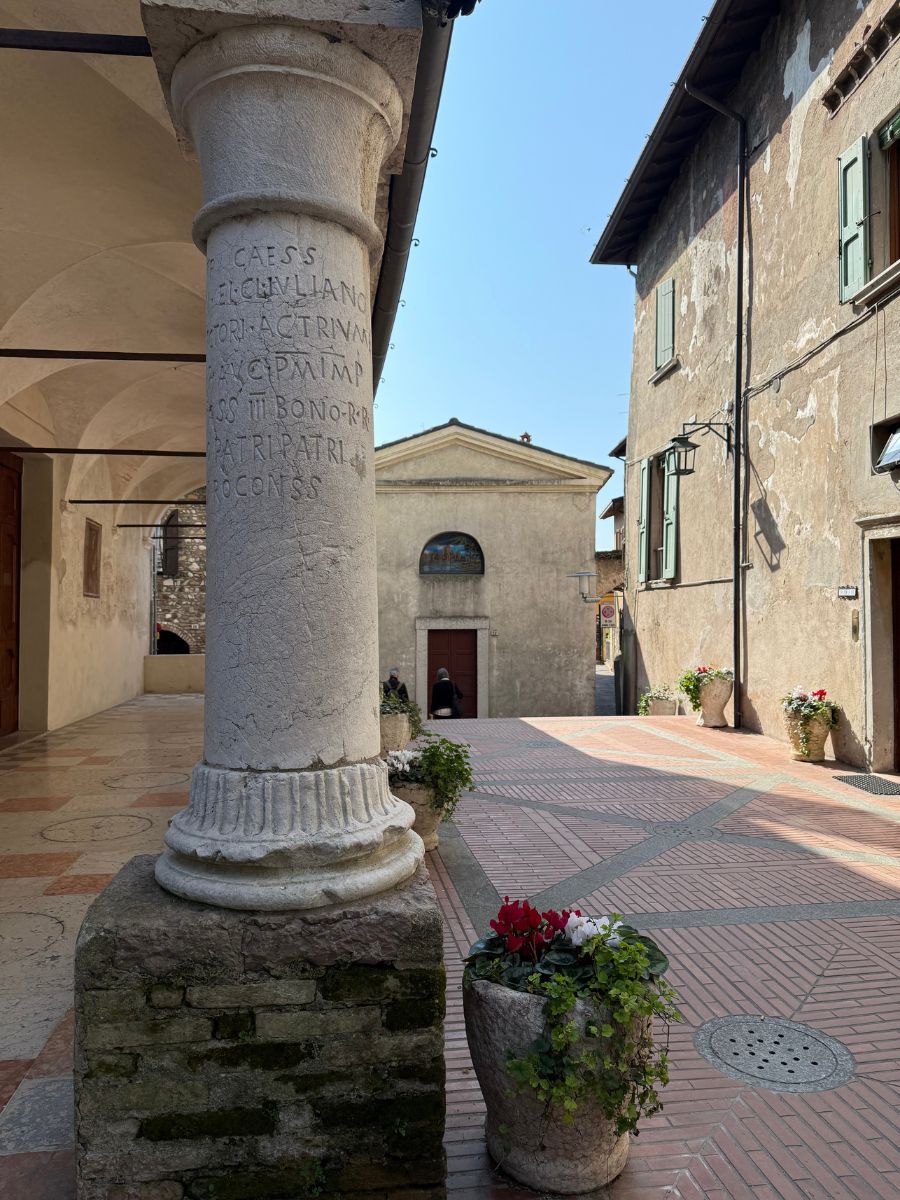 Historic column with Latin inscription in a peaceful courtyard in Sirmione, Italy, surrounded by stone buildings with shuttered windows and flower pots, under a clear blue sky.