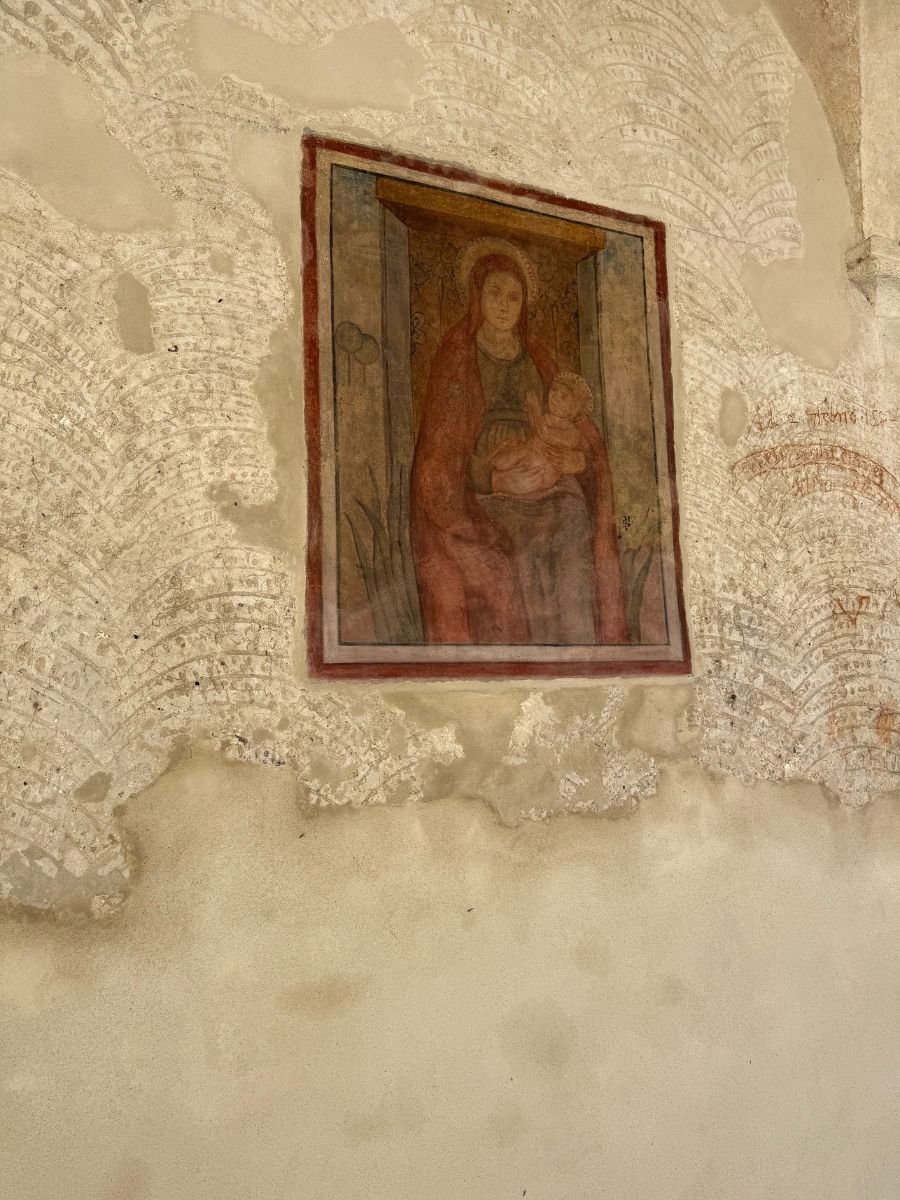 A faded fresco painting on an old plaster wall, depicting the Madonna holding the infant Jesus. The artwork is framed by a simple red border and shows signs of aging, with surrounding plaster chipped and discolored, revealing hints of earlier decorative patterns beneath.