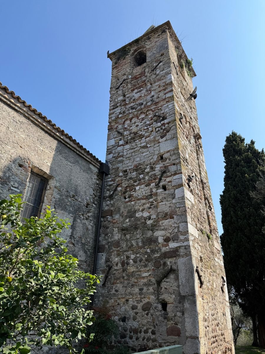 Exterior of an ancient stone bell tower in Sirmione, Italy, built with irregular stones and bricks, attached to a historic church. The structure shows signs of age with weathering and vegetation growing at the top. A lemon tree and tall cypress trees are visible nearby under a clear blue sky.