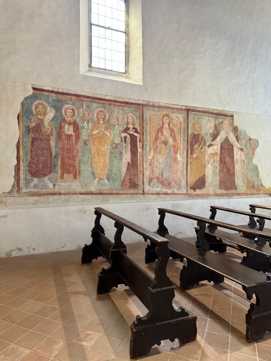 A fresco on the interior wall of a church in Sirmione, Italy, depicting a row of saints and holy figures with halos in traditional robes. The artwork, though partially faded, retains rich earth tones and is situated below a tall arched window that lets in natural light. In the foreground are dark wooden pews set on a terracotta tile floor, facing toward the fresco.