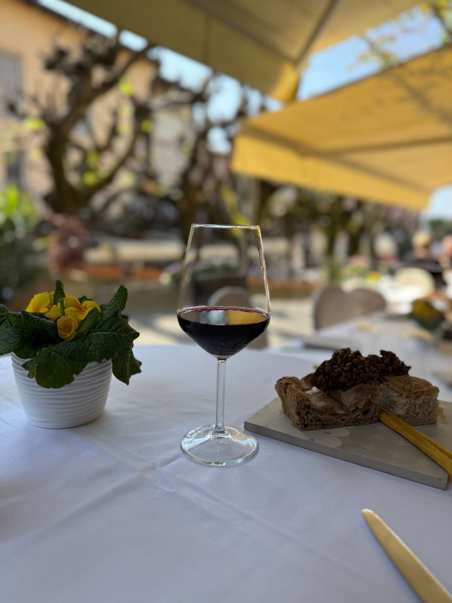 A close-up of an elegant outdoor dining table in Sirmione, Italy, set with a glass of red wine, a small plate of rustic bread topped with seeds, and a yellow flower centerpiece. The background shows blurred trees and buildings under large sunshades, evoking a relaxed and sunny café atmosphere.