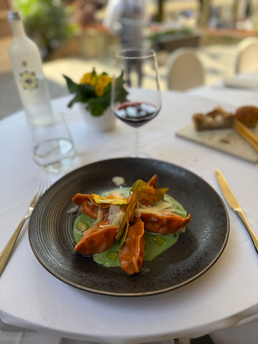 A gourmet dish of stuffed pasta arranged on a dark ceramic plate, served over a green sauce and garnished with zucchini blossoms and cheese shavings. The table is set outdoors at a restaurant in Sirmione, Italy, with a glass of red wine, a bottle of water, and fresh bread in the background, creating a refined and inviting dining atmosphere.