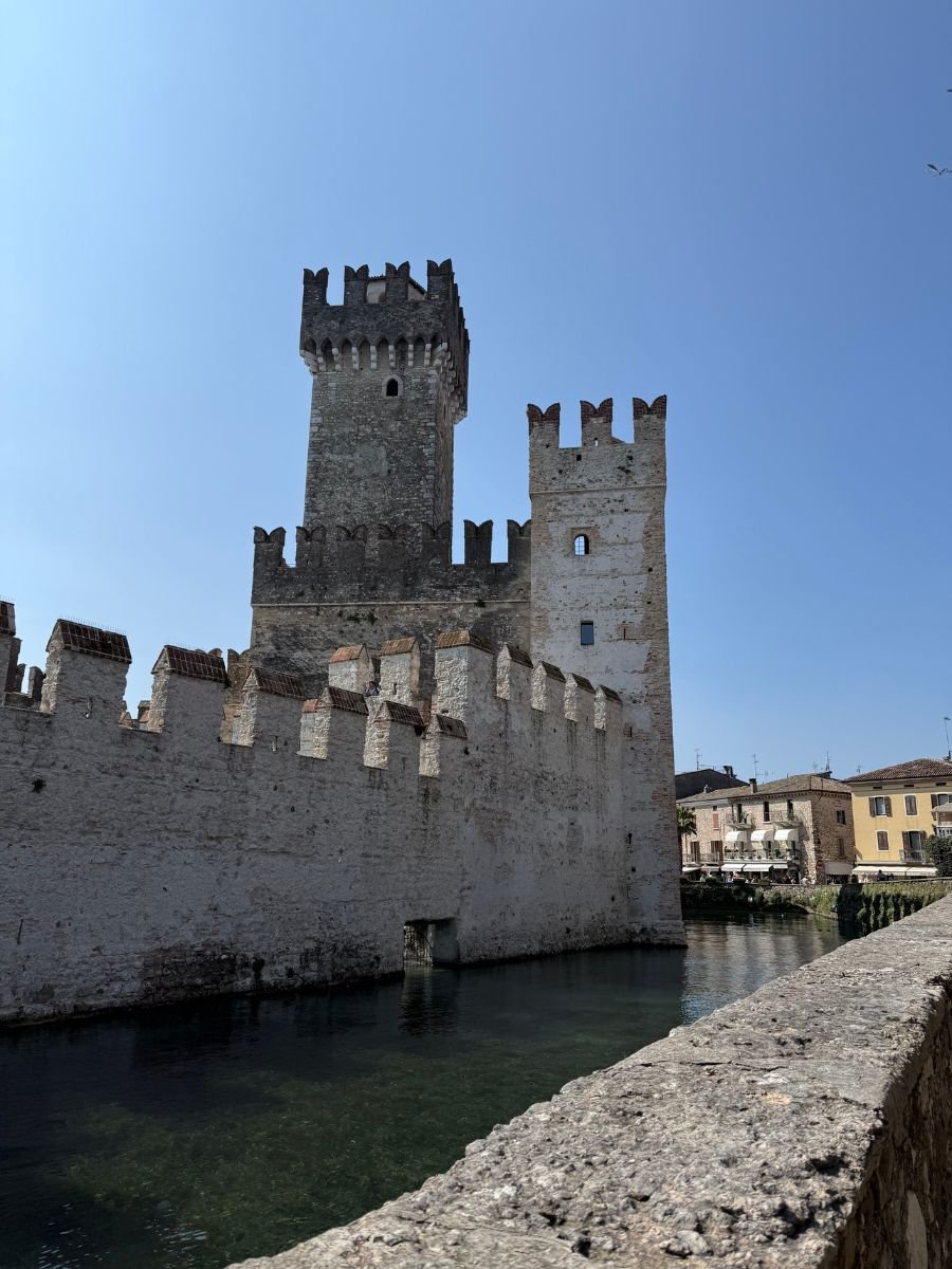 A view of Scaliger Castle in Sirmione, Italy, showing its stone walls and tall crenellated towers reflected in the still waters of the surrounding moat. The sky is clear and blue, and a few buildings from the old town are visible in the background to the right.