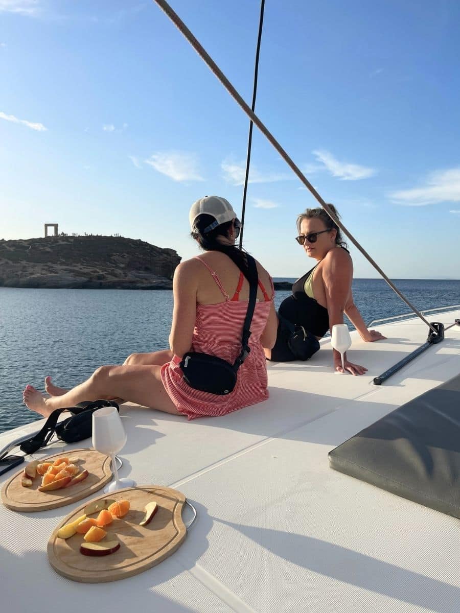 Two women sitting on a boat deck, enjoying the ocean view near a rocky island with a historic archway. Wooden plates with sliced fruit and white glasses are on the deck in the foreground.