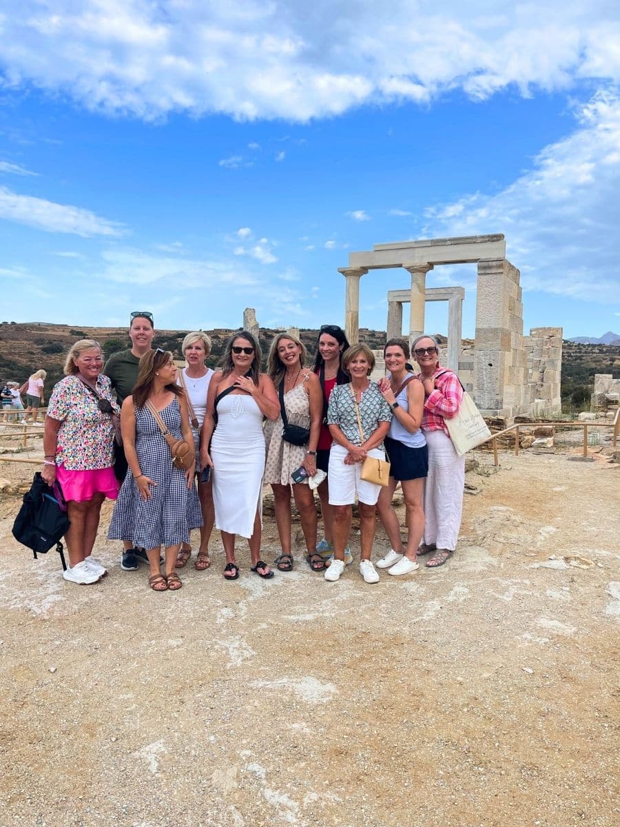 A group photo of nine people standing in front of ancient ruins, with clear blue skies and a rocky landscape. The ruins feature a prominent stone doorway and columns.
