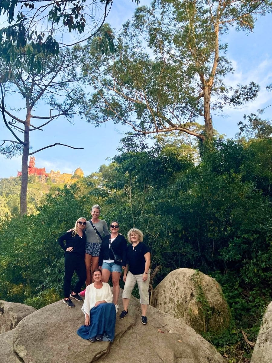 Five women posing on large rocks surrounded by lush greenery, with a colorful castle visible in the distance atop a hill under a clear blue sky.