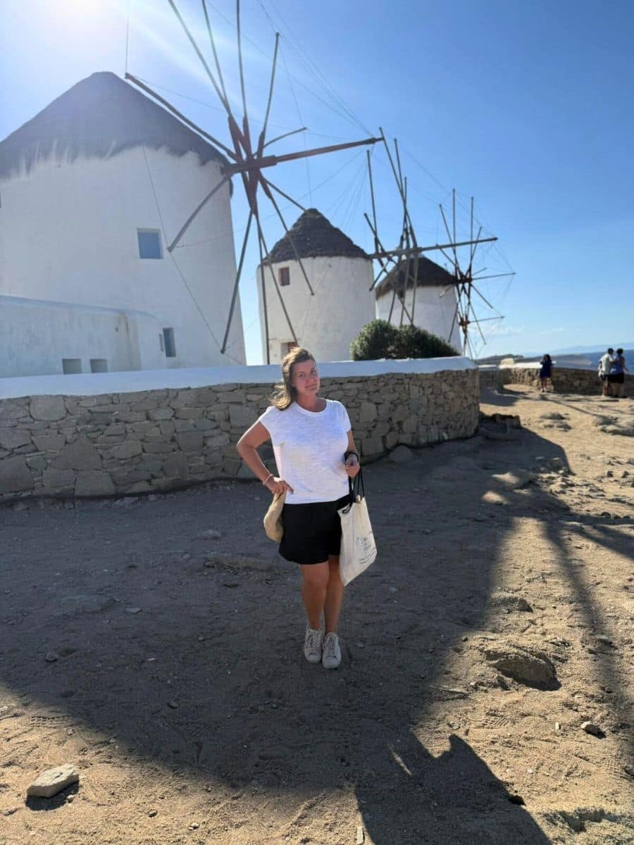 A woman stands confidently in front of traditional whitewashed windmills with thatched roofs, set against a clear blue sky. The shadow of the windmill's blades stretches across the sandy ground, adding depth to the picturesque scene.