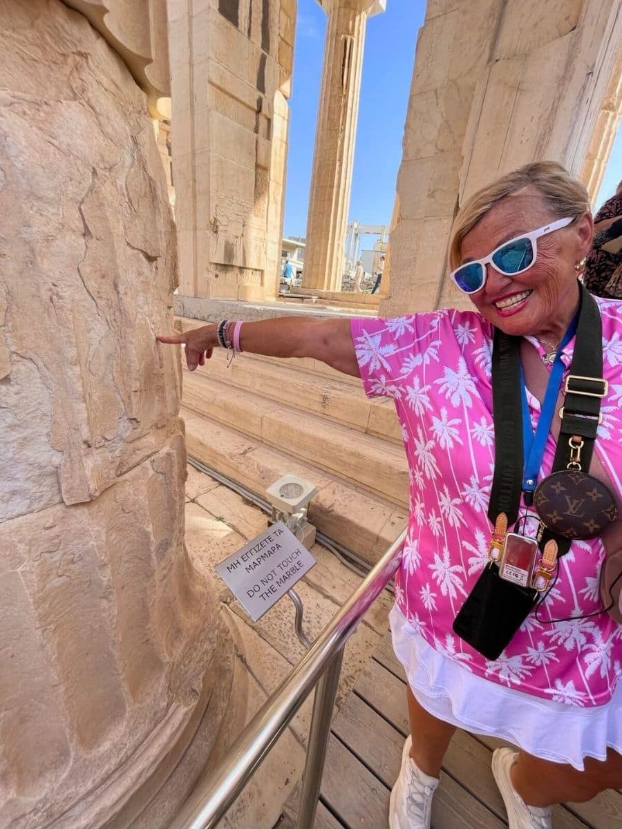 A visitor humorously points at a sign reading 'DO NOT TOUCH THE MARBLE' while standing near an ancient column. The bright pink tropical shirt and playful expression add a lively contrast to the historical surroundings of the ancient structure.