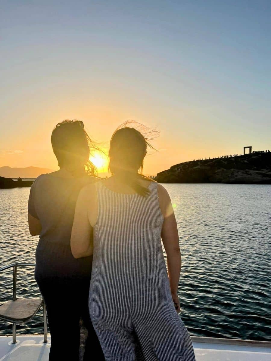 Two women stand side by side on a boat, gazing at a breathtaking sunset over the water. The sun casts a golden glow, illuminating the rippling waves and a distant hillside adorned with the silhouette of an ancient structure.