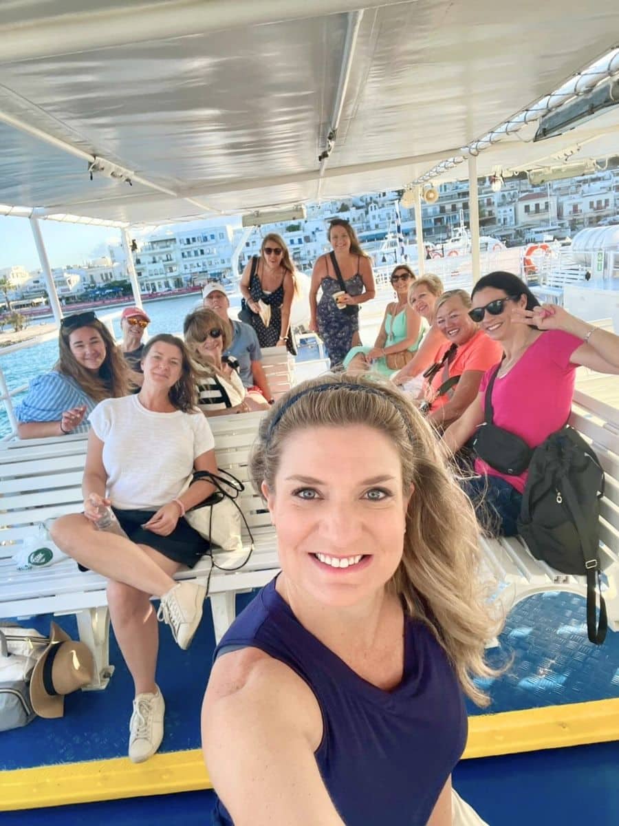 A lively group photo taken on a boat with a backdrop of a picturesque coastal town. Everyone is smiling and enjoying the moment under the shaded deck, radiating joy and camaraderie.