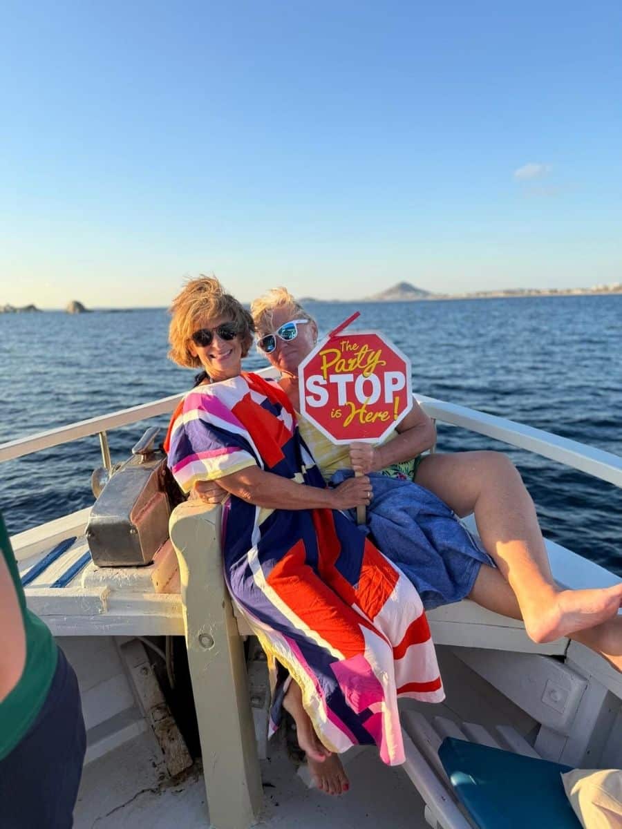 Two people sitting on a boat, smiling and holding a sign that reads 'The Party STOP is Here!' They are wrapped in colorful clothing, enjoying a sunny day with calm ocean waters and distant landforms in the background.