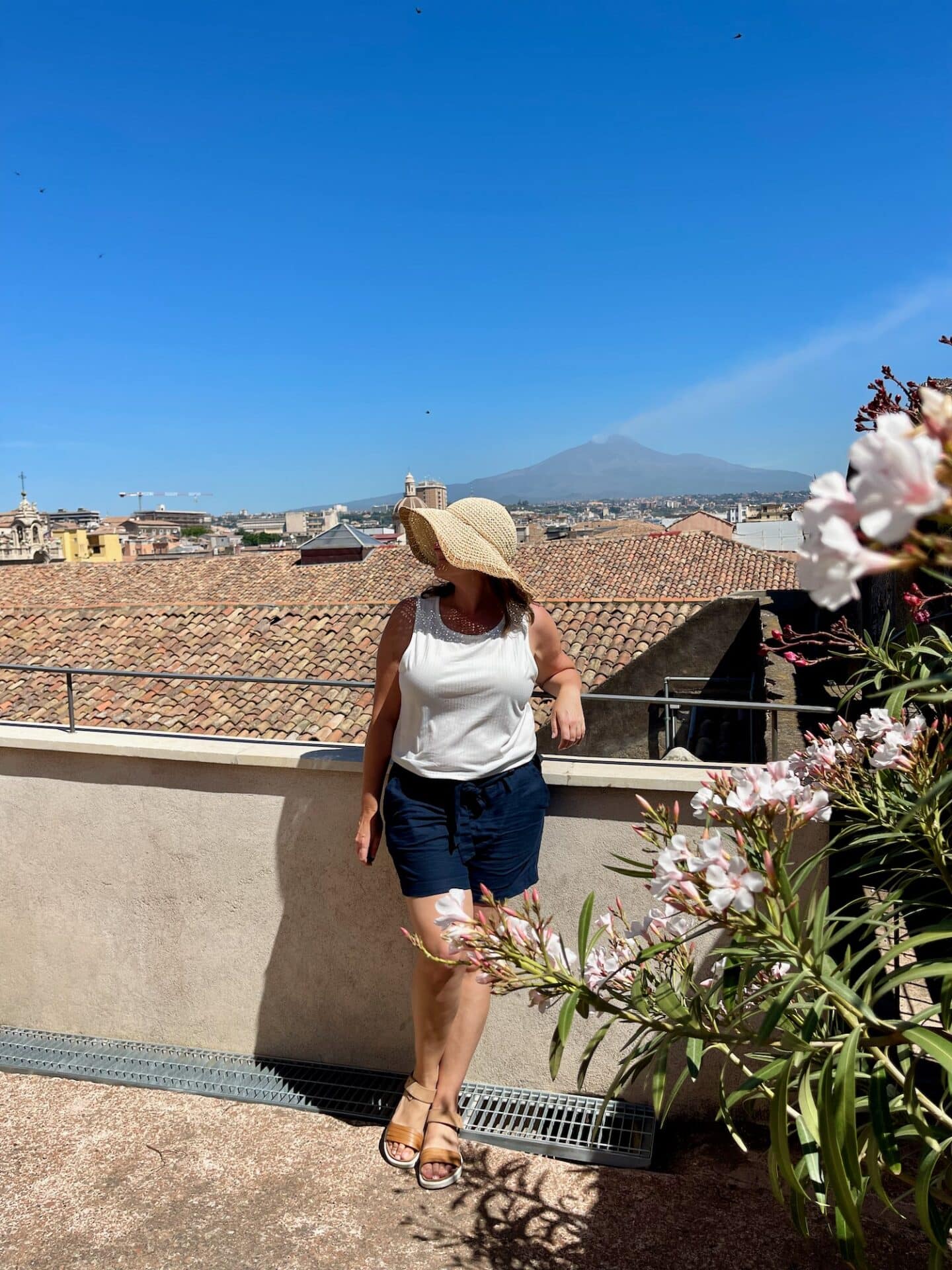 A woman in a sunhat posing on a rooftop terrace in Catania with Mount Etna in the background.
