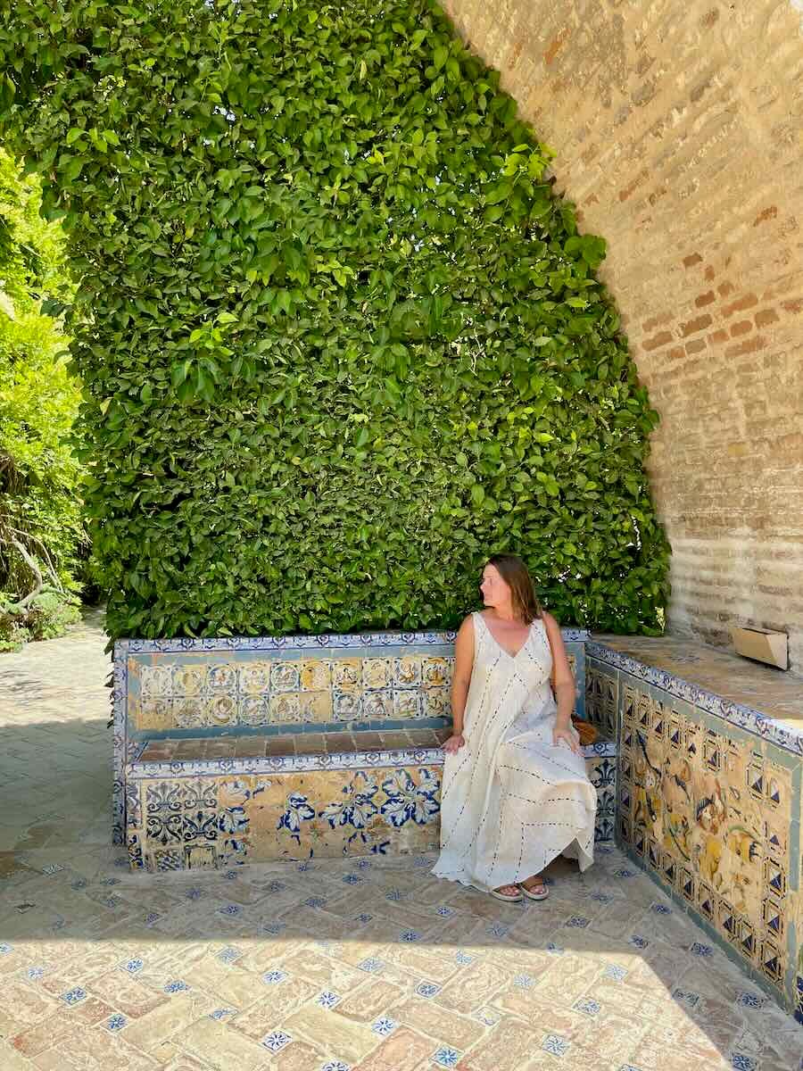 A woman is sitting alone on a beautifully tiled bench surrounded by lush green foliage within the gardens of Real Alcázar in Seville. The image captures the tranquility and detailed artistry of the surroundings.