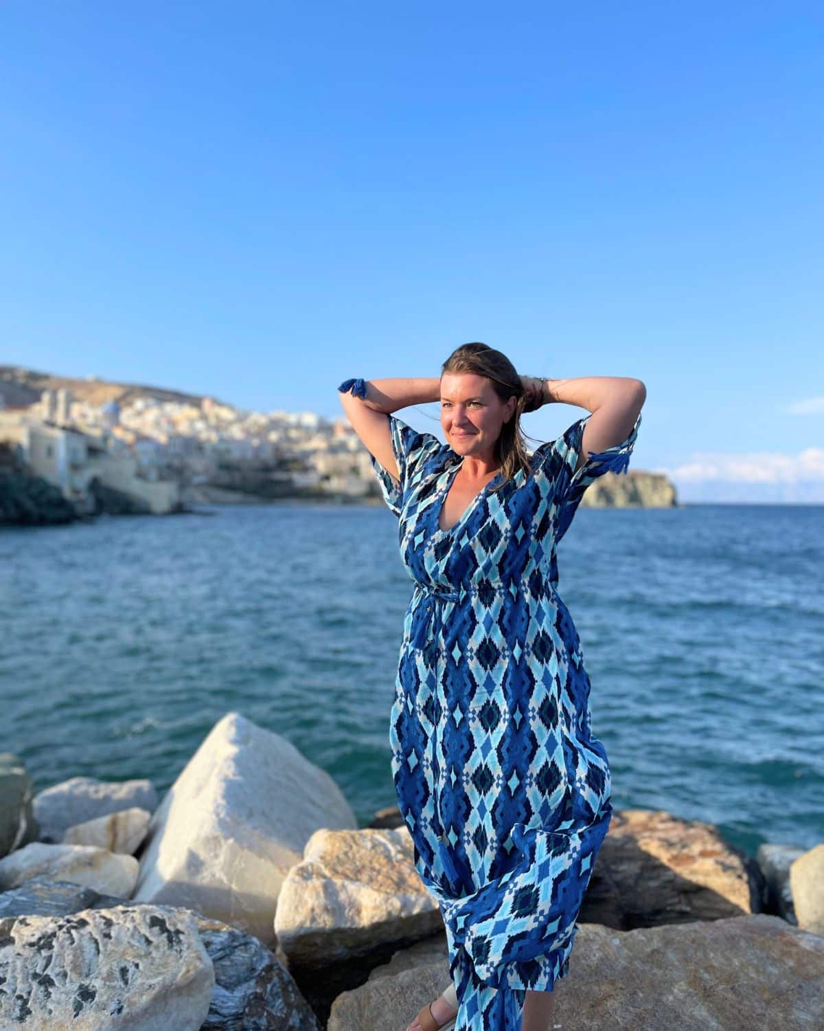 A woman in a white dress stands in front of a historical stone archway, with a blurred dome structure visible in the background. Her expression is serene and she's partially shielding her face from the sun with her hand. The image may evoke the sense of peaceful solitude one might experience while traveling alone.