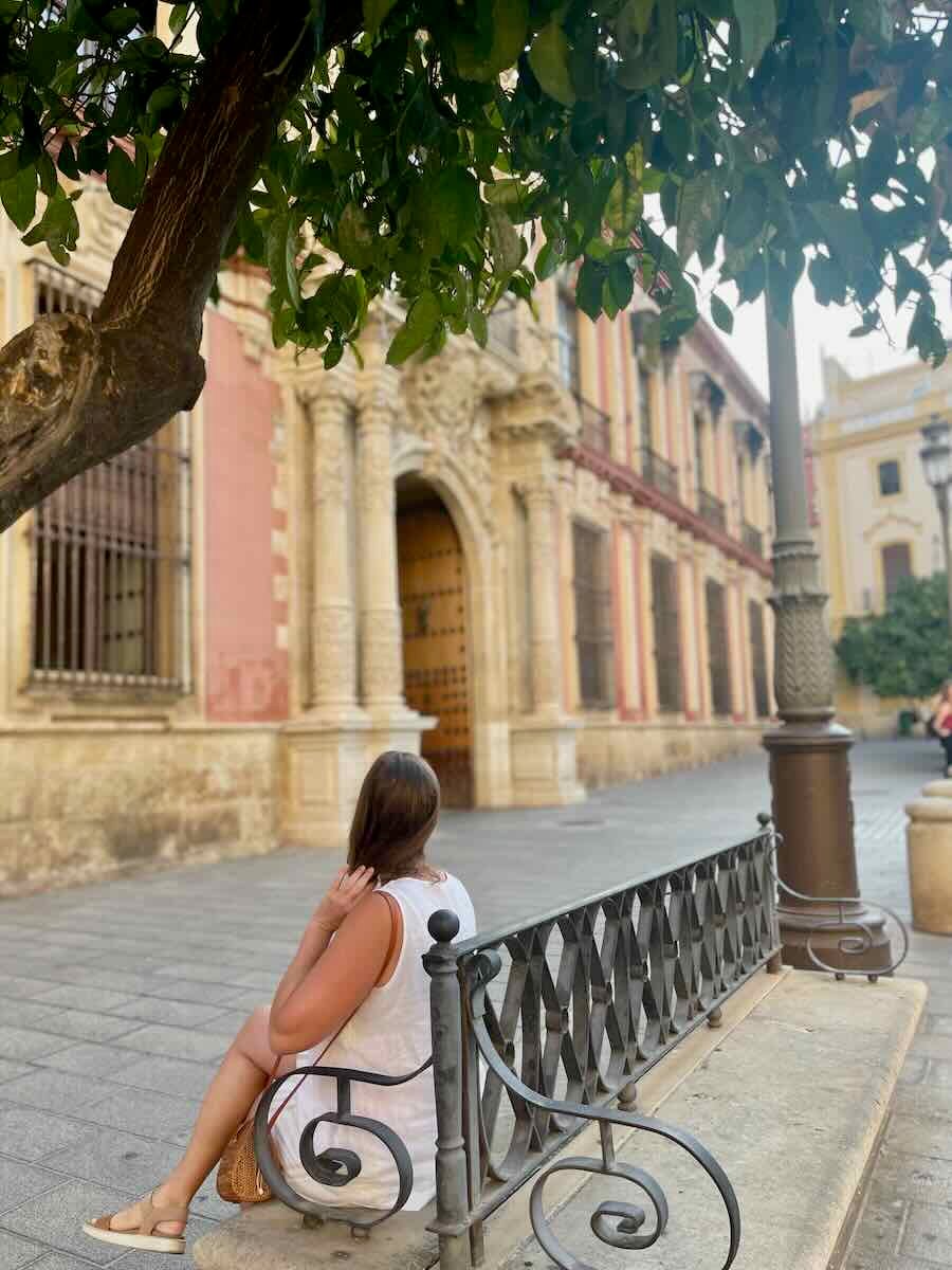The image shows a woman sitting on a bench under the shade of a tree, with historic architecture in the background. This seems to be a quiet, reflective moment in a charming Seville street.