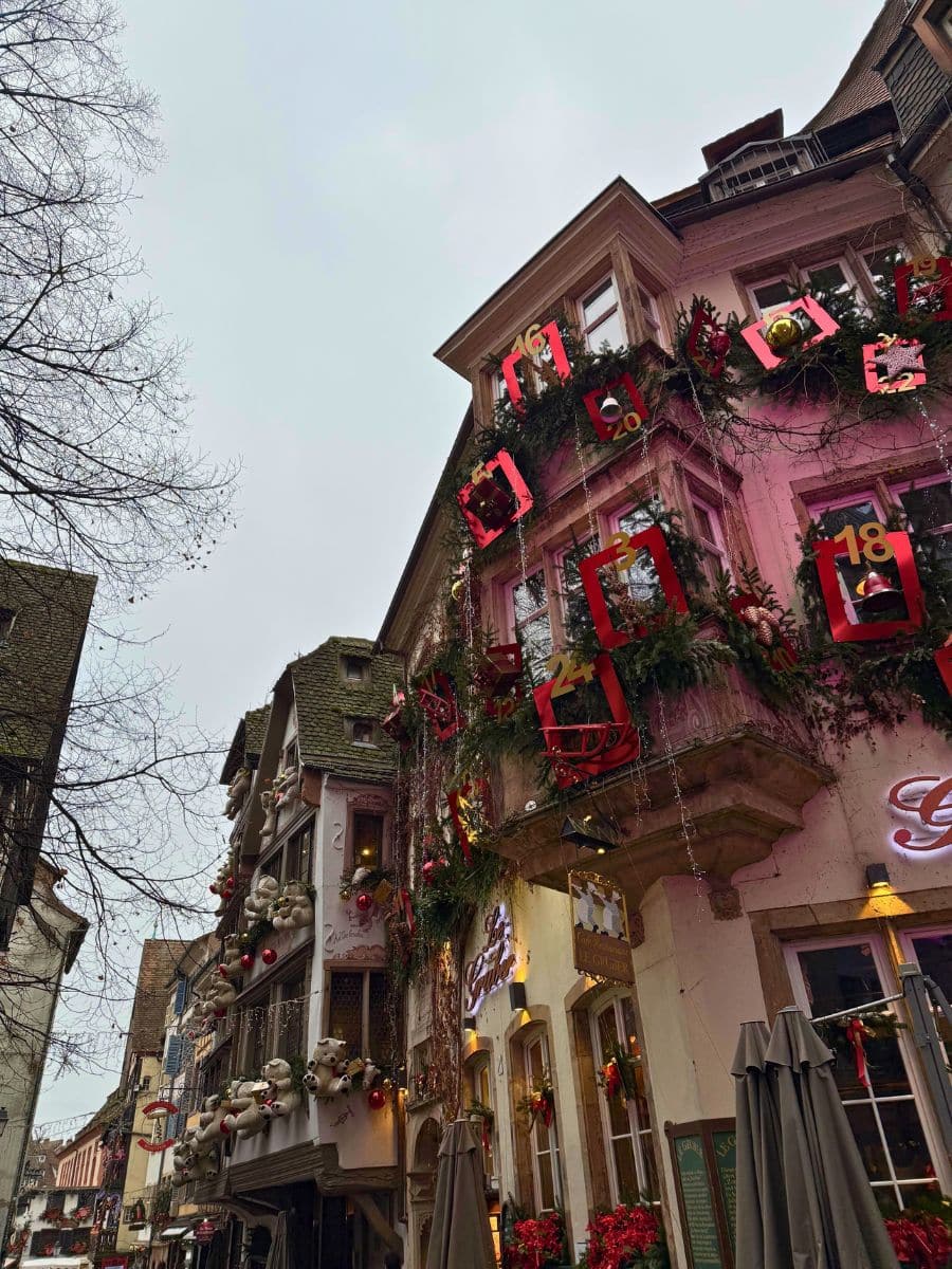 A beautifully decorated building in Strasbourg with festive garlands and an advent calendar theme, featuring red-framed numbers on the windows. Adjacent buildings are adorned with holiday decorations, including teddy bears and ornaments. The cobblestone street and the intricate architecture add to the enchanting Christmas atmosphere.