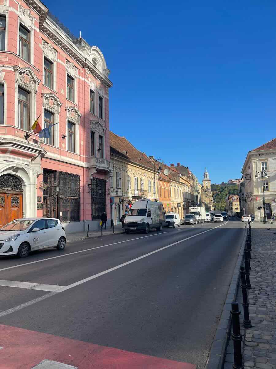 This image features a row of colorful buildings along a quiet street in Bra?ov, Romania. The street is lined with pastel-hued buildings, including a prominent pink building on the left. The bright blue sky contrasts with the historic architecture, and cars are visible along the road.