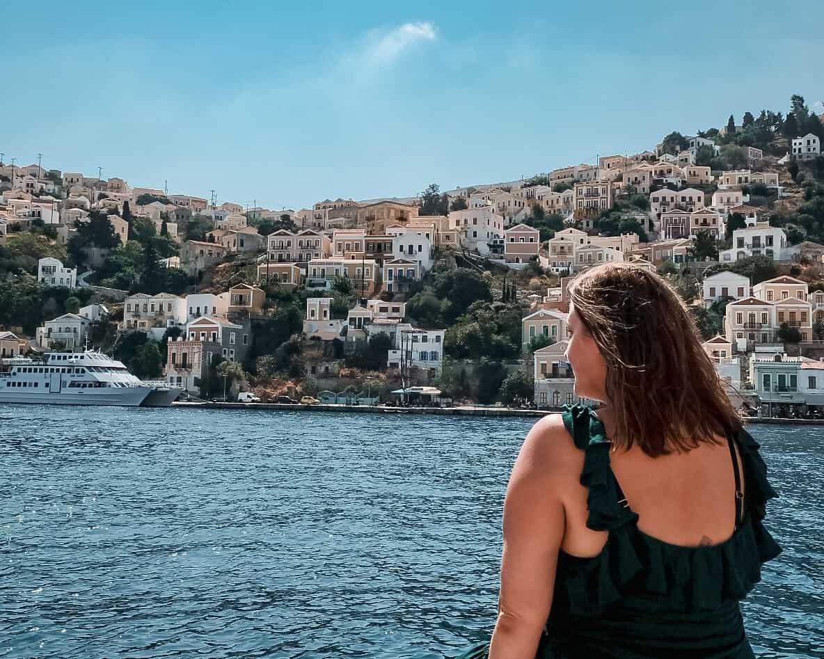 A woman in a green ruffled dress is gazing towards a beautiful hillside town by the sea. The town is densely packed with classic Mediterranean-style houses in shades of white and pastel, cascading down the hill to the water's edge where boats are docked. The sea is a deep blue and the sky is clear, adding to the idyllic coastal ambiance of the scene.