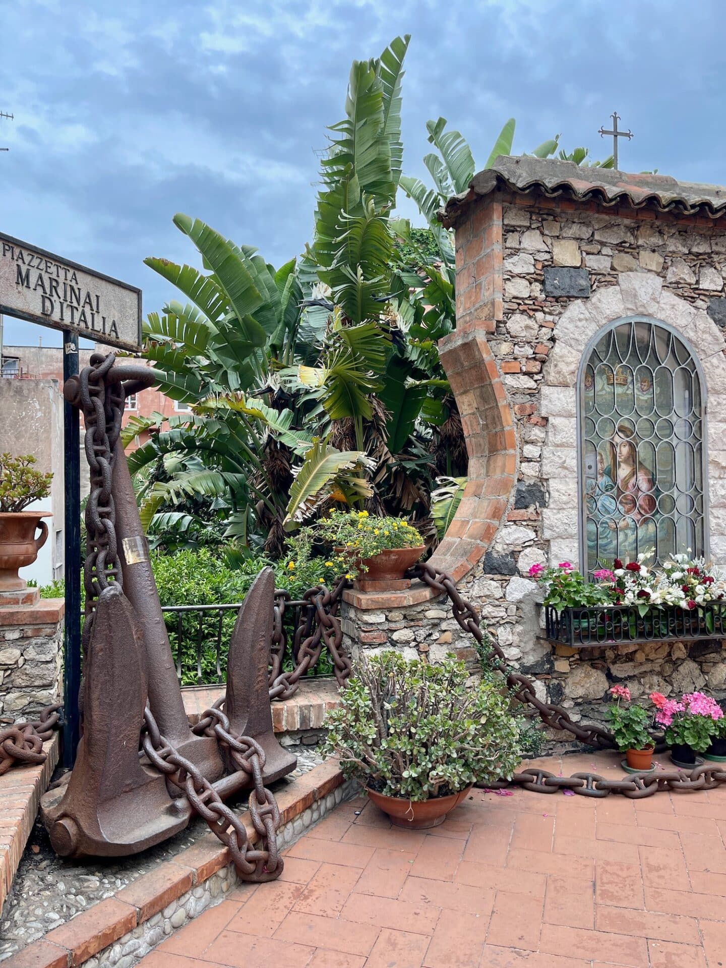 A corner in Taromina featuring a large maritime anchor with chains, lush greenery, and a stone structure with a decorated window.