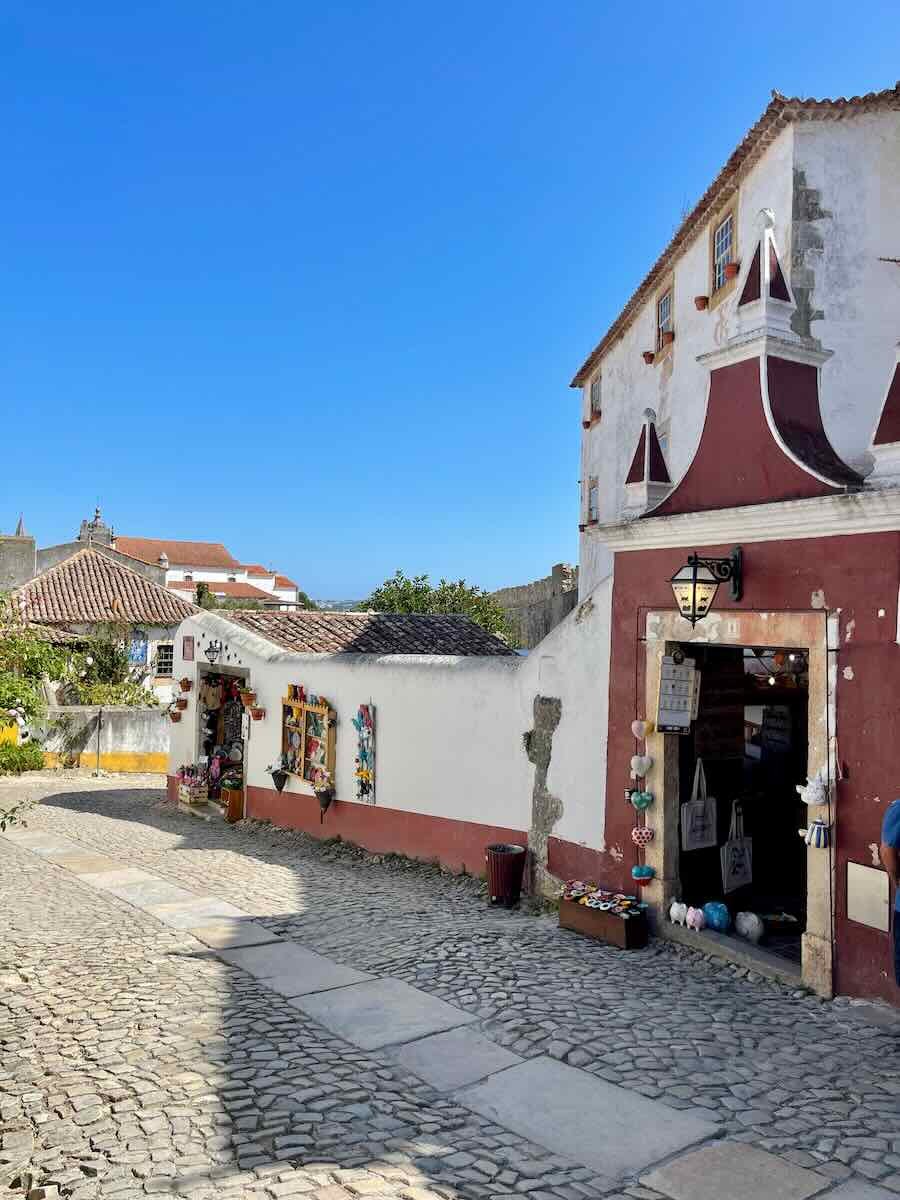 A quaint cobblestone street in Óbidos, lined with small traditional houses, one of which has a rustic red facade with a decorative gable. Souvenir shops add color and vibrancy to this medieval town.