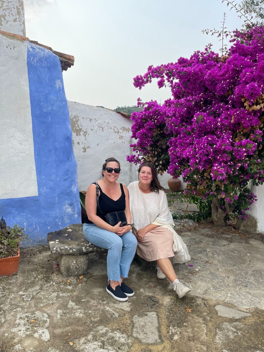 Two women sitting outdoors on a stone bench, one wearing sunglasses, a black top, and jeans, the other in a light, flowy dress. They're surrounded by rustic walls, one painted white and blue, and a vibrant purple bougainvillea tree blooms beside them. The setting appears serene and Mediterranean.