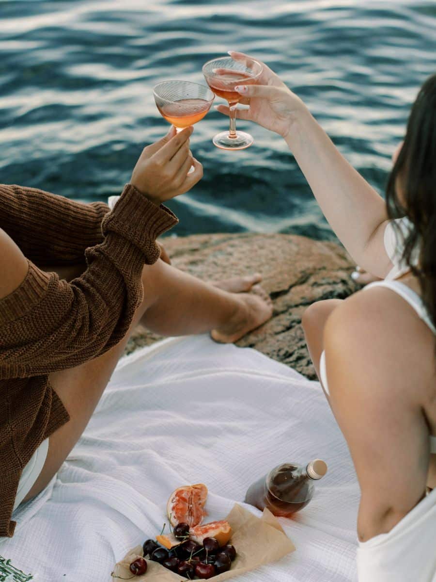 Two women toast with glasses of rosé by the water, enjoying a scenic seaside picnic with fresh fruit and drinks on a white blanket. A perfect moment of relaxation, connection, and travel memories.