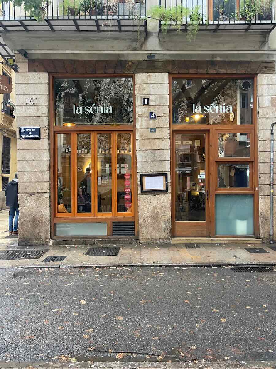 The front of La Senia restaurant in Valencia, featuring a stone facade with large wooden-framed windows and doors. A sign with the restaurant’s name is visible above the entrance, set against a wet, cobblestone street.