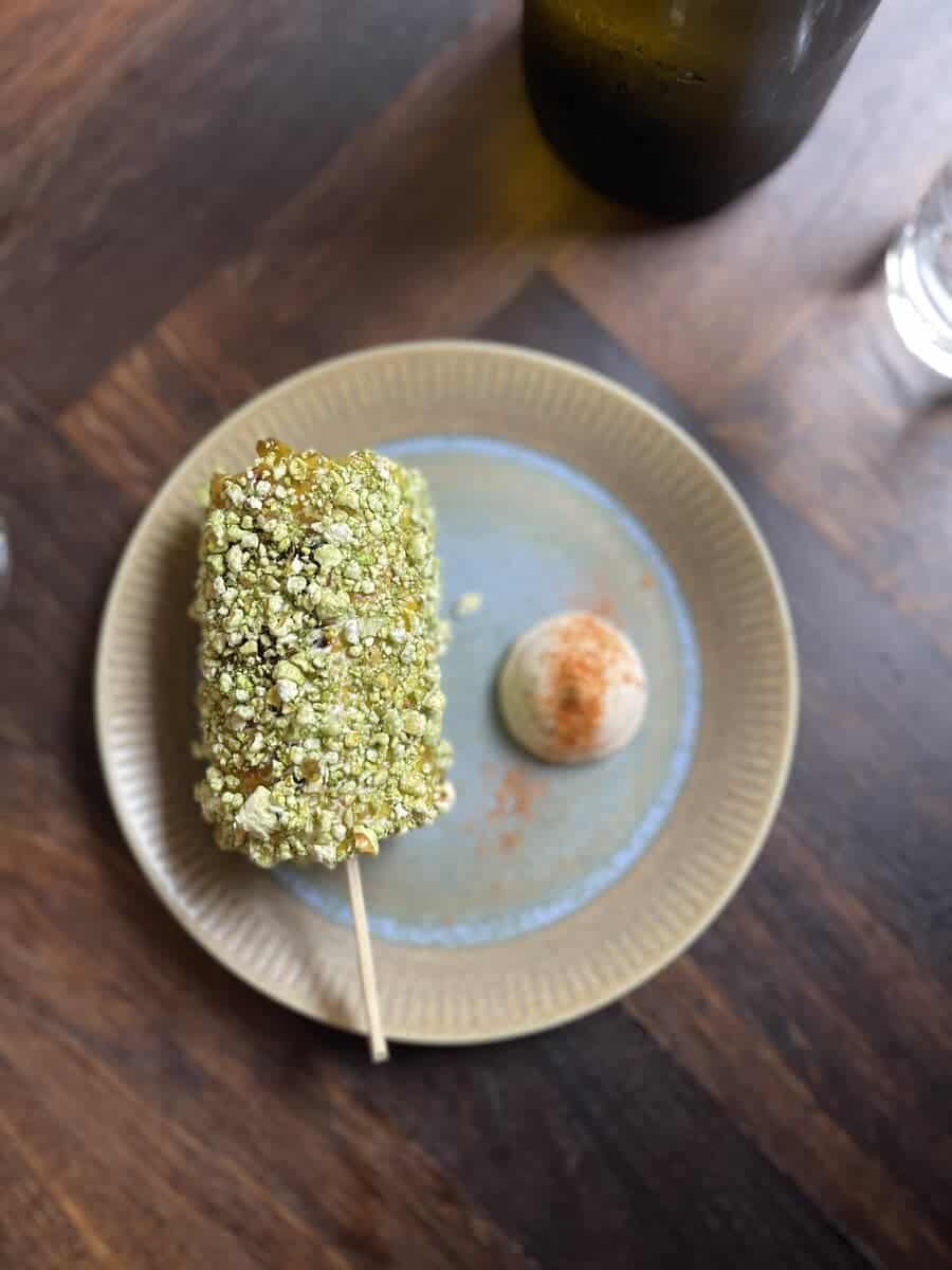 A gourmet dish featuring a pistachio-crusted dessert on a stick, served with a scoop of seasoned cream, presented on a ceramic plate in a Copenhagen restaurant.
