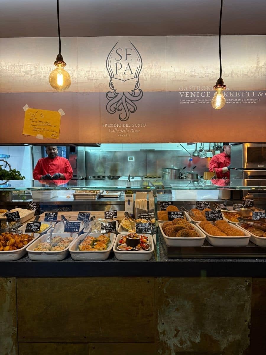 A Venetian deli counter at SEPA in Calle della Bissa, displaying a variety of freshly prepared local dishes like polpette, seafood, and vegetables. Warm lighting and chefs in red uniforms add to the inviting, authentic atmosphere.