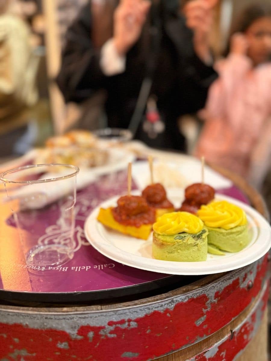 A plate of Venetian street food resting on a rustic red barrel at SEPA in Calle della Bissa, featuring colorful savory bites including meatballs in tomato sauce and mini vegetable flans topped with bright yellow purée. Diners enjoy their meals in the lively background.