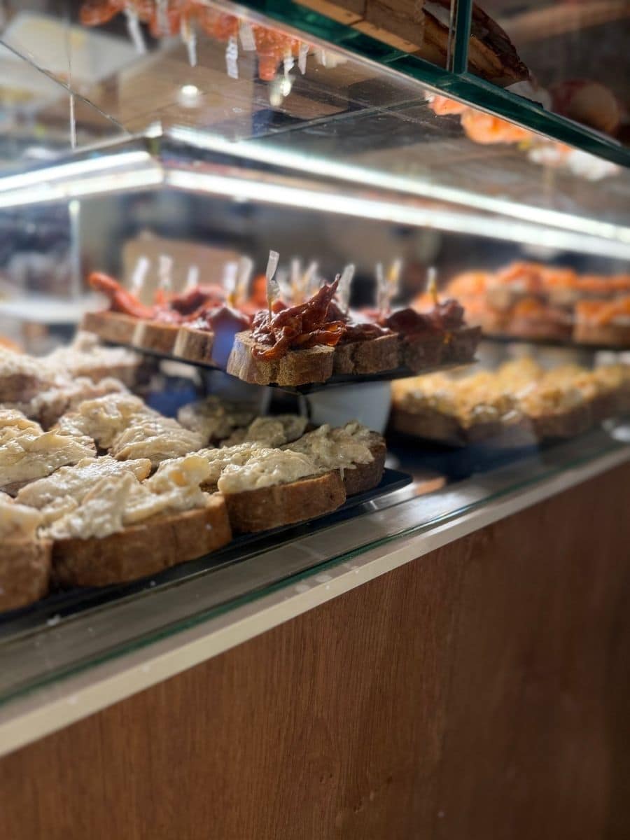 Close-up of traditional Venetian cicchetti — small open-faced sandwiches topped with creamy spreads and sun-dried tomatoes, displayed in a glass counter.