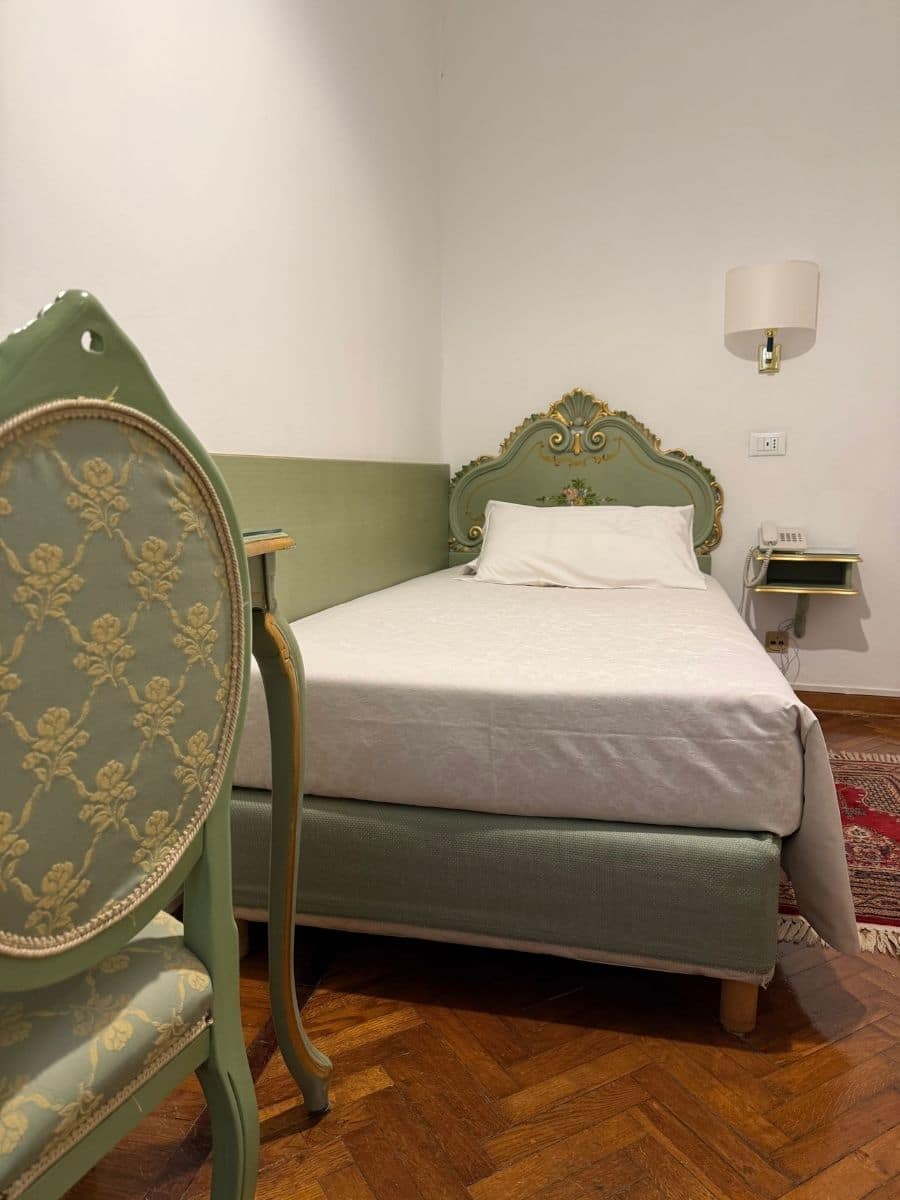 A vintage-style single bed in a Venetian hotel room, featuring an ornate green headboard with floral details, a matching upholstered chair, and a polished wooden floor with a red accent rug.
