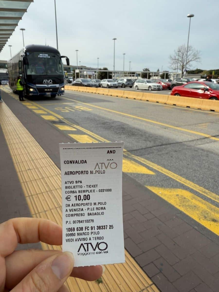 Close-up of a hand holding an ATVO bus ticket from Marco Polo Airport to Piazzale Roma in Venice, with a black ATVO airport shuttle bus approaching in the background.
