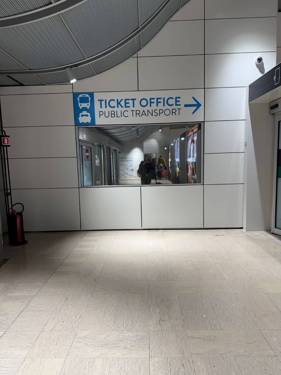 A blue and white sign on a tiled wall indicates the direction to the "TICKET OFFICE PUBLIC TRANSPORT" with a right-pointing arrow and icons of a bus and train. The entrance to the office is visible through a window, showing people inside.