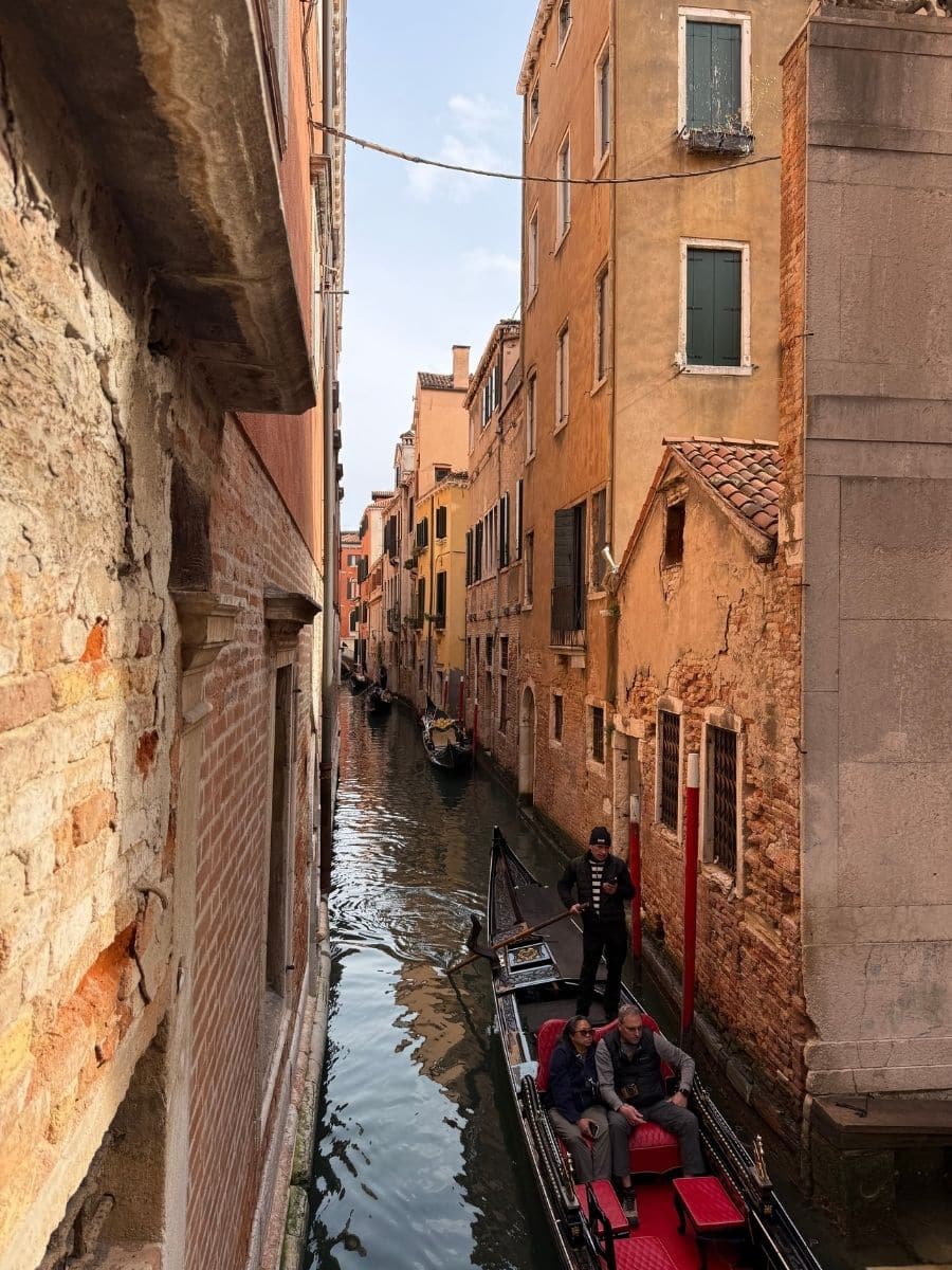 A view looking down into a narrow canal in Venice, Italy. A black gondola with red seating carries two passengers and is being propelled by a gondolier. Tall, aged buildings in shades of orange and beige line both sides of the waterway, rising directly from the water.