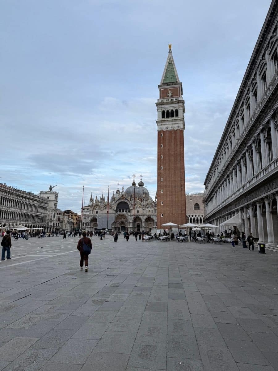 The iconic St. Mark's Square in Venice, captured in a wide view that encompasses the grandeur of St. Mark's Basilica, the height of the Campanile, and the surrounding historic architecture. People are seen strolling and gathering in the vast piazza, experiencing one of Venice's most famous locations.
