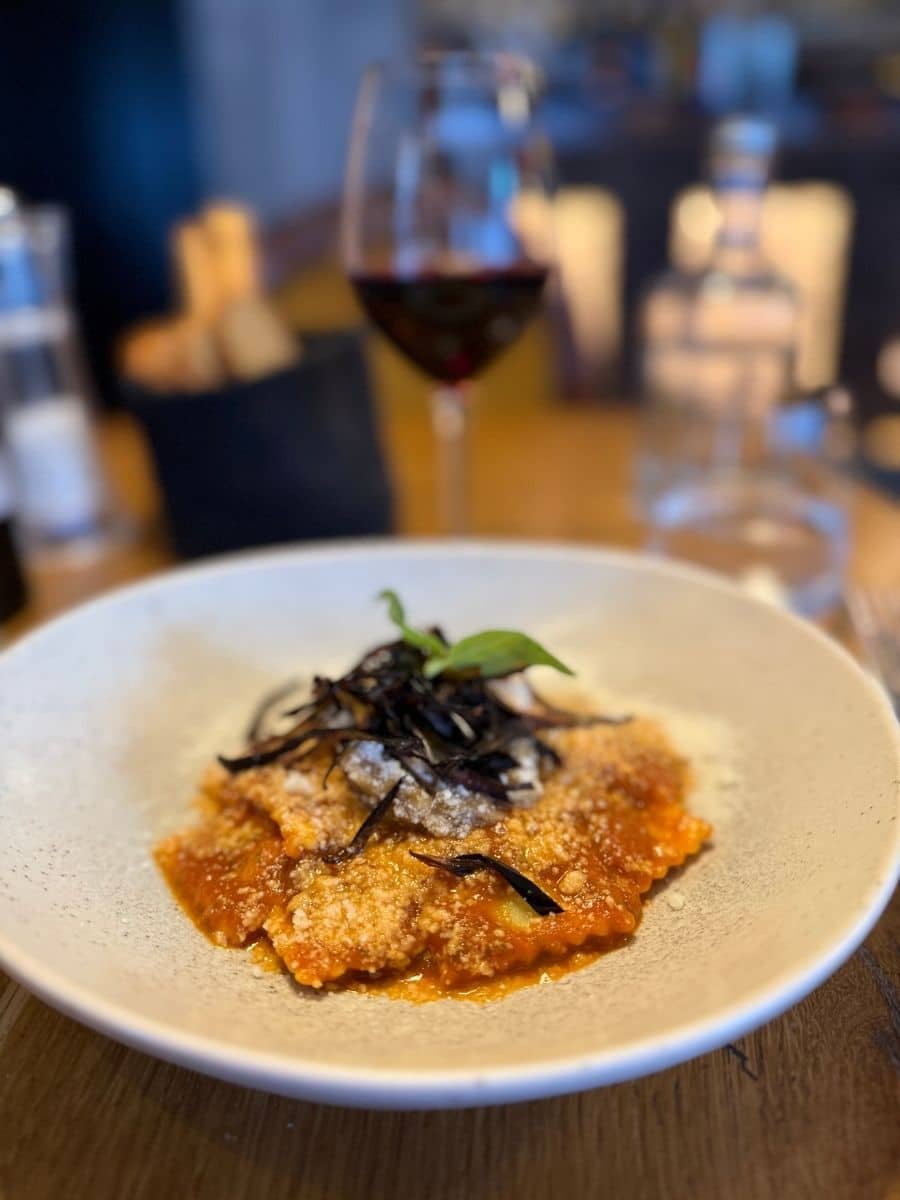 A close-up of a white, textured plate featuring several ravioli filled with a light-colored filling and covered in a rich red tomato sauce. The dish is garnished with grated cheese, dark, crispy fried leeks or onions, and a few small green basil leaves. A wine glass filled with red wine is visible in the blurred background, suggesting a restaurant setting.