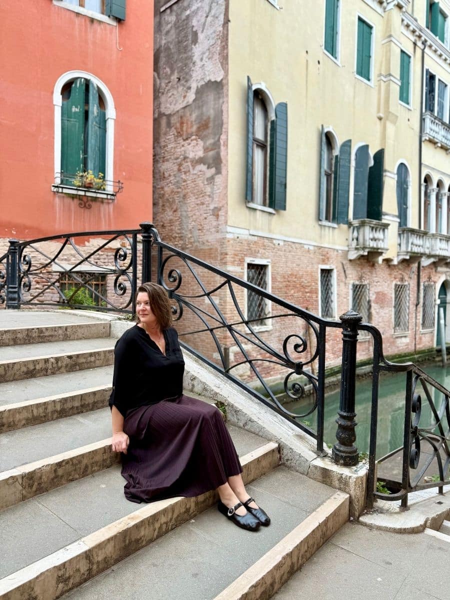 A peaceful moment in Venice, where Melissa is seated on the steps of a charming bridge, taking in the sights of the surrounding canals and historic buildings. The vibrant colors of the buildings and the gentle flow of the water create a classic Venetian atmosphere.