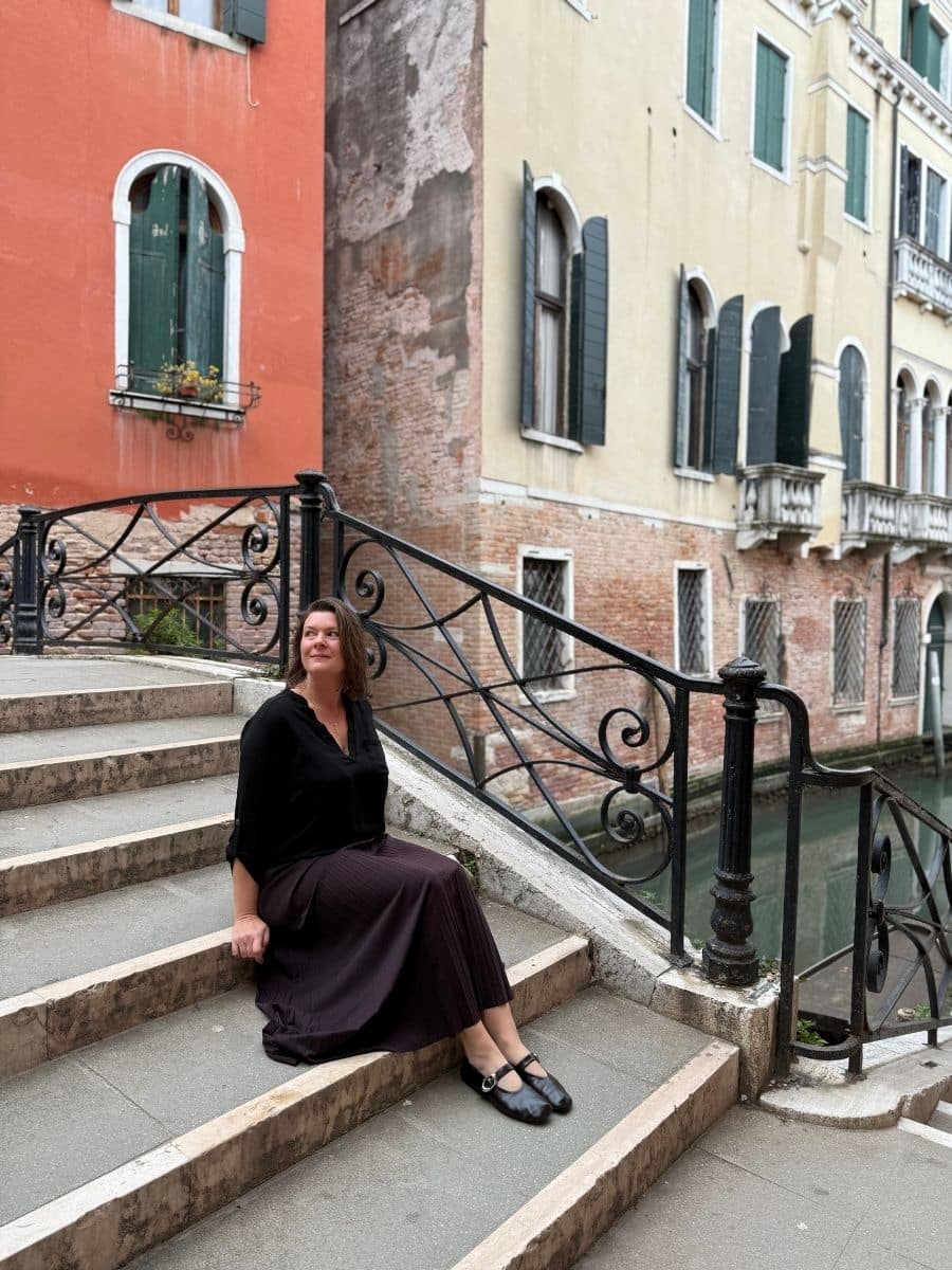 Melissa wearing a black top and a dark skirt sits on the stone steps of a pedestrian bridge that crosses a canal in Venice, Italy. Ornate black metal railings line the bridge. Colorful, aged buildings with window shutters flank the canal. The water is visible below the bridge.