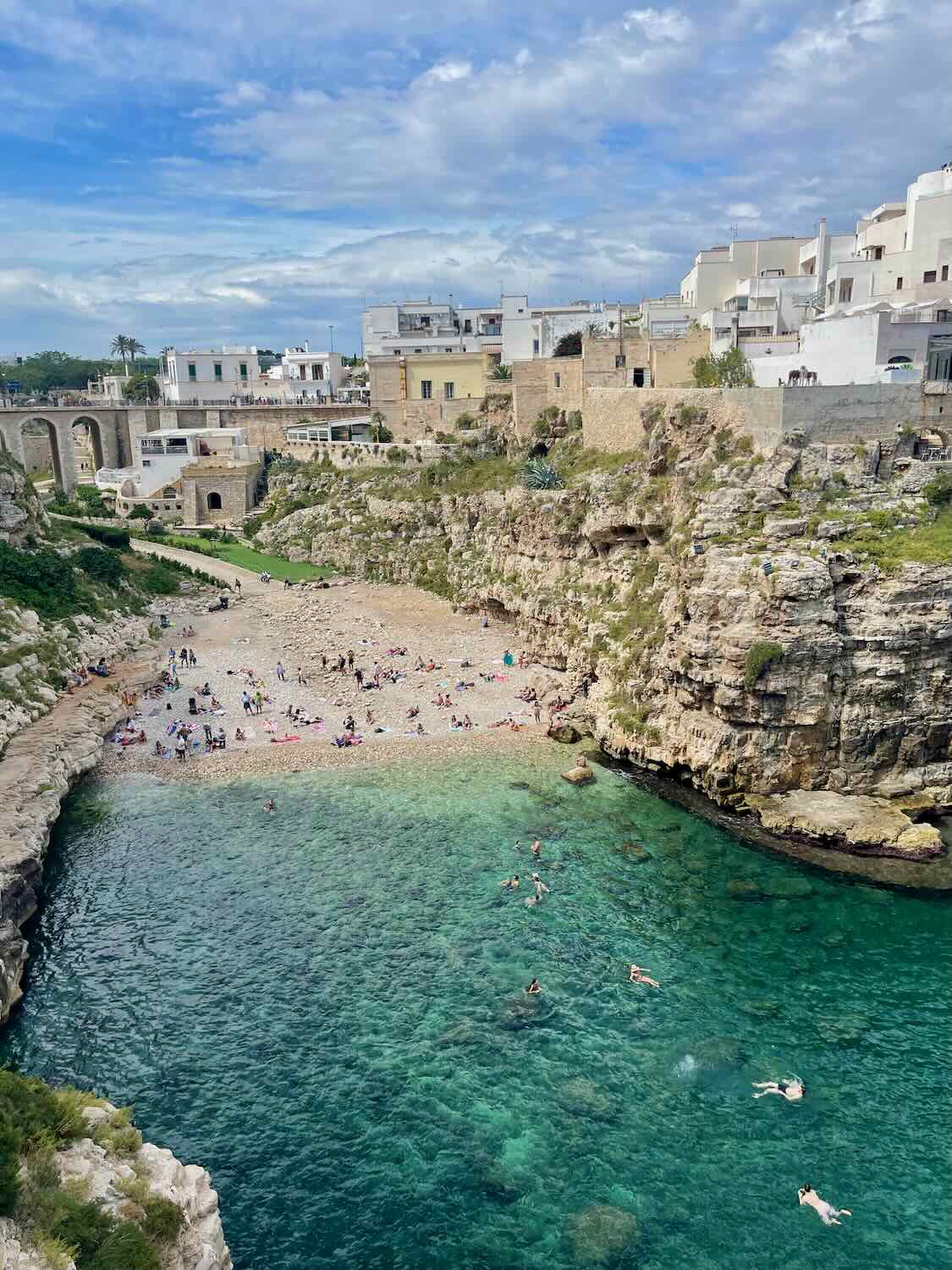 This image depicts a beautiful coastal scene, with a small cove featuring clear turquoise water and a beach nestled between rocky cliffs. The beach is populated with sunbathers, and the surrounding area has traditional white and beige buildings, likely part of a historic town. The overall atmosphere suggests a Mediterranean coastal location, perfect for swimming and relaxation.