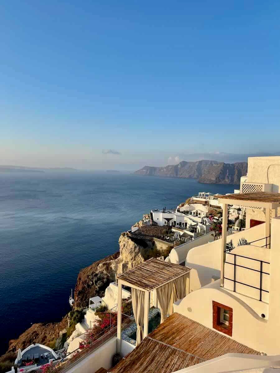 View of the Santorini coastline with white-washed buildings perched on a cliff overlooking the blue Aegean Sea, with distant mountains under a clear sky.