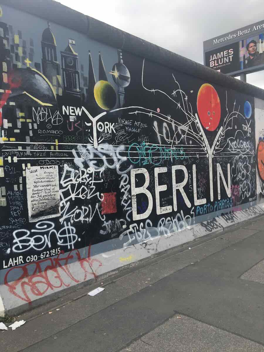A section of the Berlin Wall with colorful graffiti and a mural featuring the word 'Berlin' in large letters. The mural is a mix of cityscape and abstract elements, representing Berlin's vibrant street art culture.