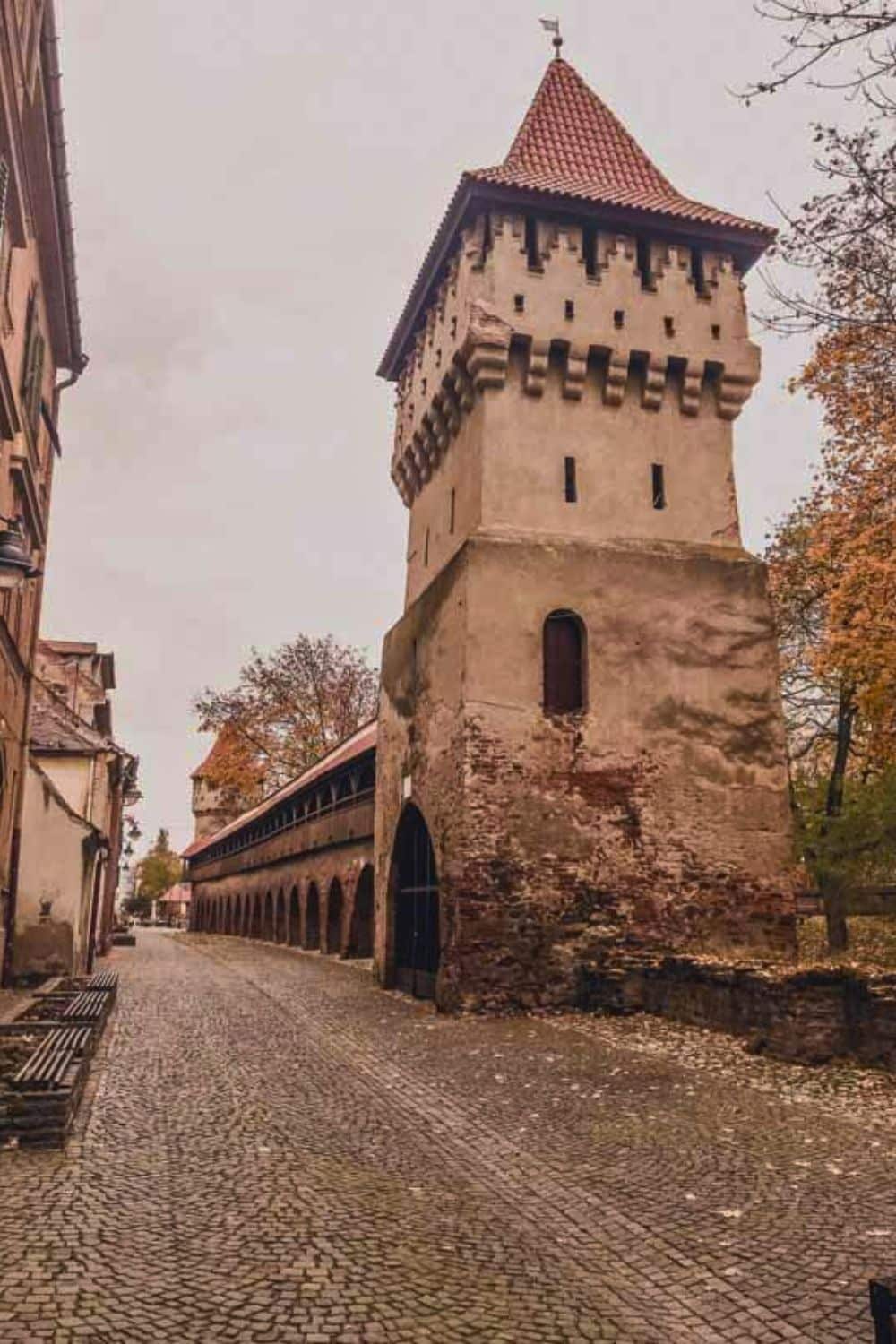 An old defensive tower with a textured facade and conical roof stands beside a covered wooden gallery, part of a historic fortification along a deserted cobblestone street