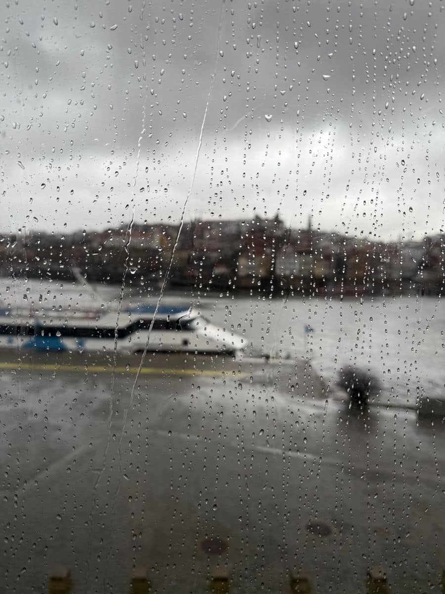 A rainy day view through a window covered in raindrops. In the background, a river, a docked white and blue boat, and a cityscape with buildings on a hill are visible under a gray, overcast sky. A blurred figure walks along the wet pavement.