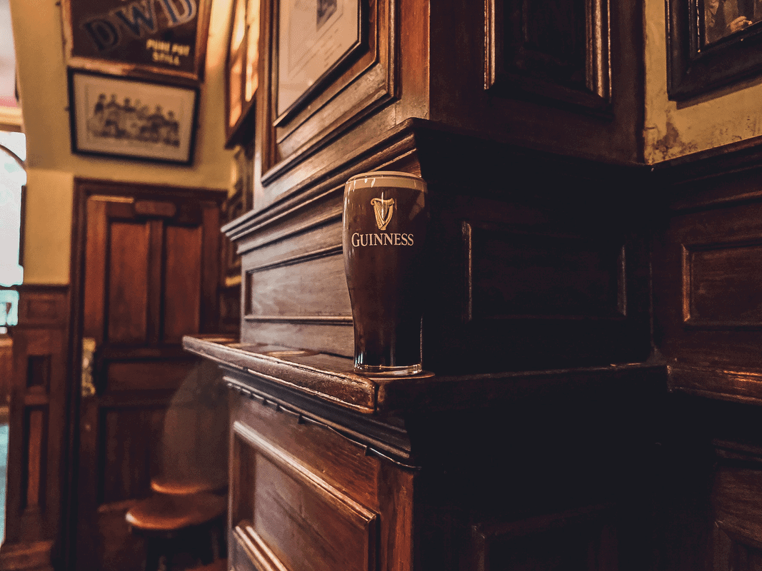 A pint of Guinness stout rests on the polished wooden ledge of an Irish pub with warm ambient lighting, framed by vintage framed pictures and traditional wood-paneled walls.