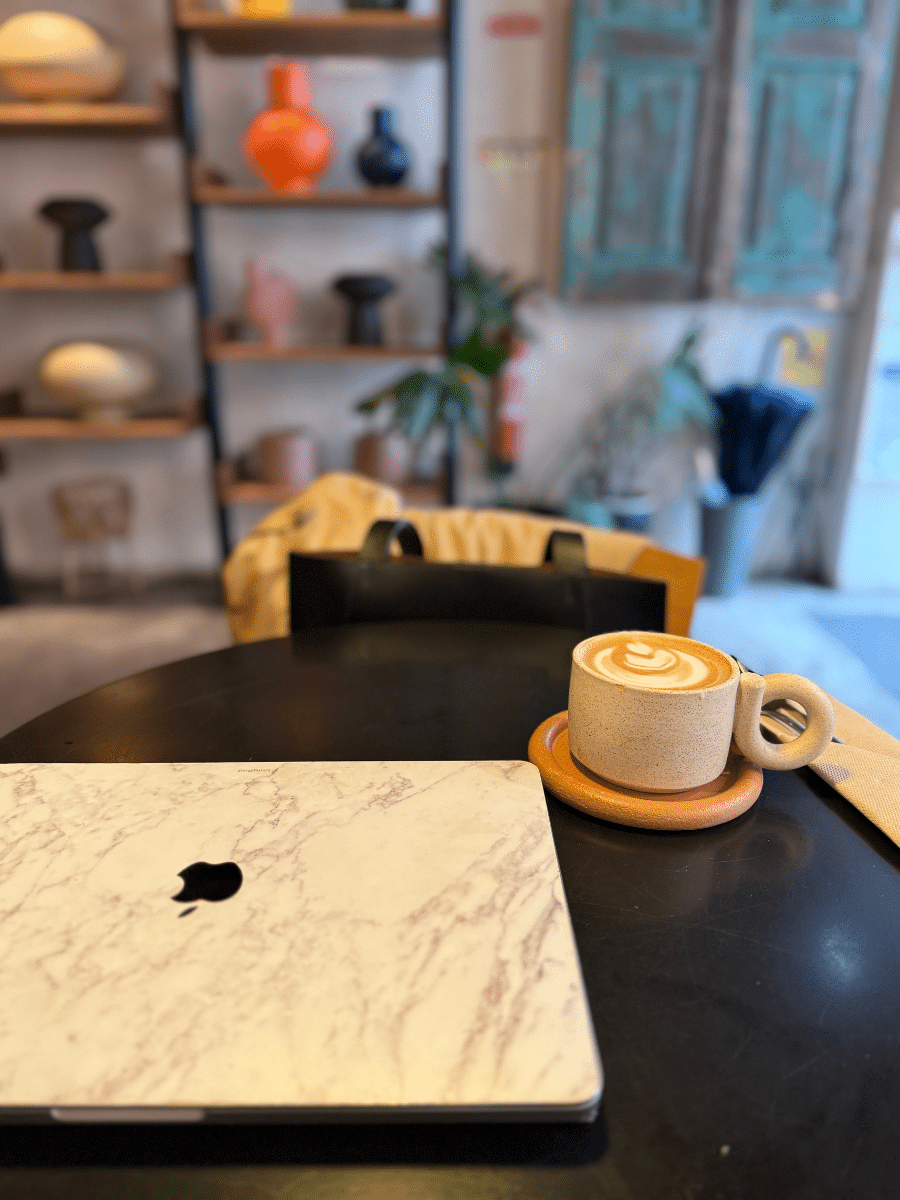 A cozy workspace setup at Marquise Cafe, featuring a marble-patterned laptop on a sleek black table. Beside it, a creamy latte served in a unique ceramic cup with a circular handle, placed on a matching saucer. The background includes stylish shelves with colorful decor pieces and rustic elements.