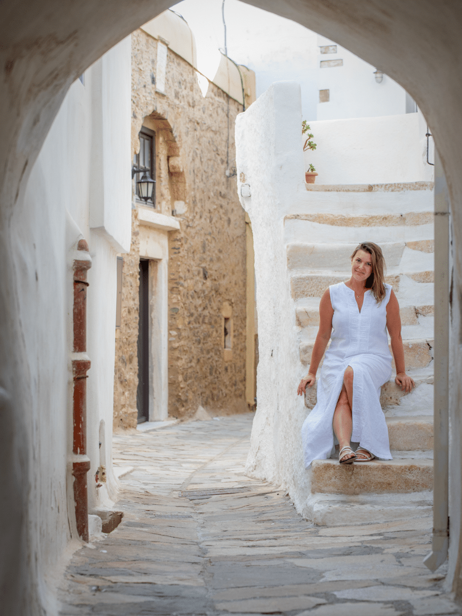 Solo female traveler in a white dress sitting on stone steps in a narrow, charming alleyway with traditional whitewashed buildings in Naxos, Greece.