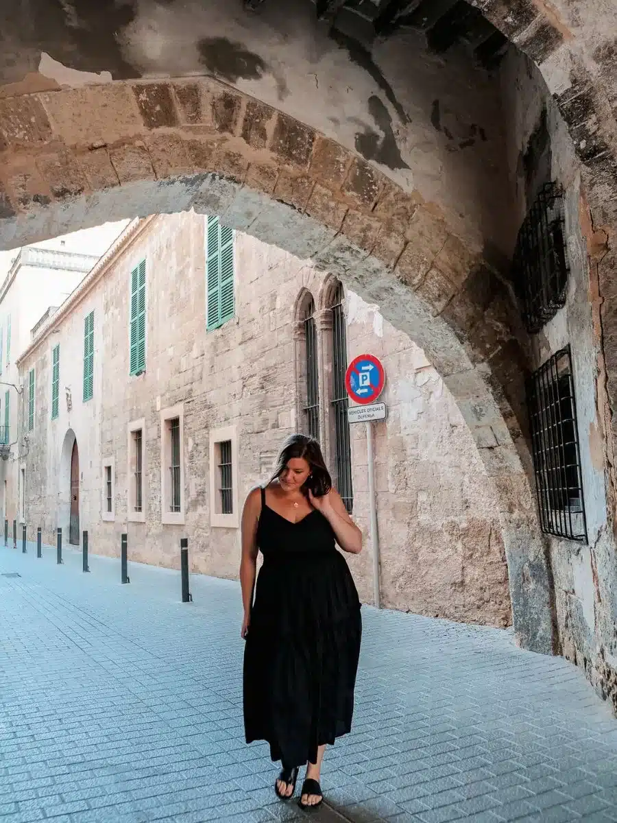A woman in a black sleeveless dress walks under a stone archway in a narrow street lined with historic beige buildings.