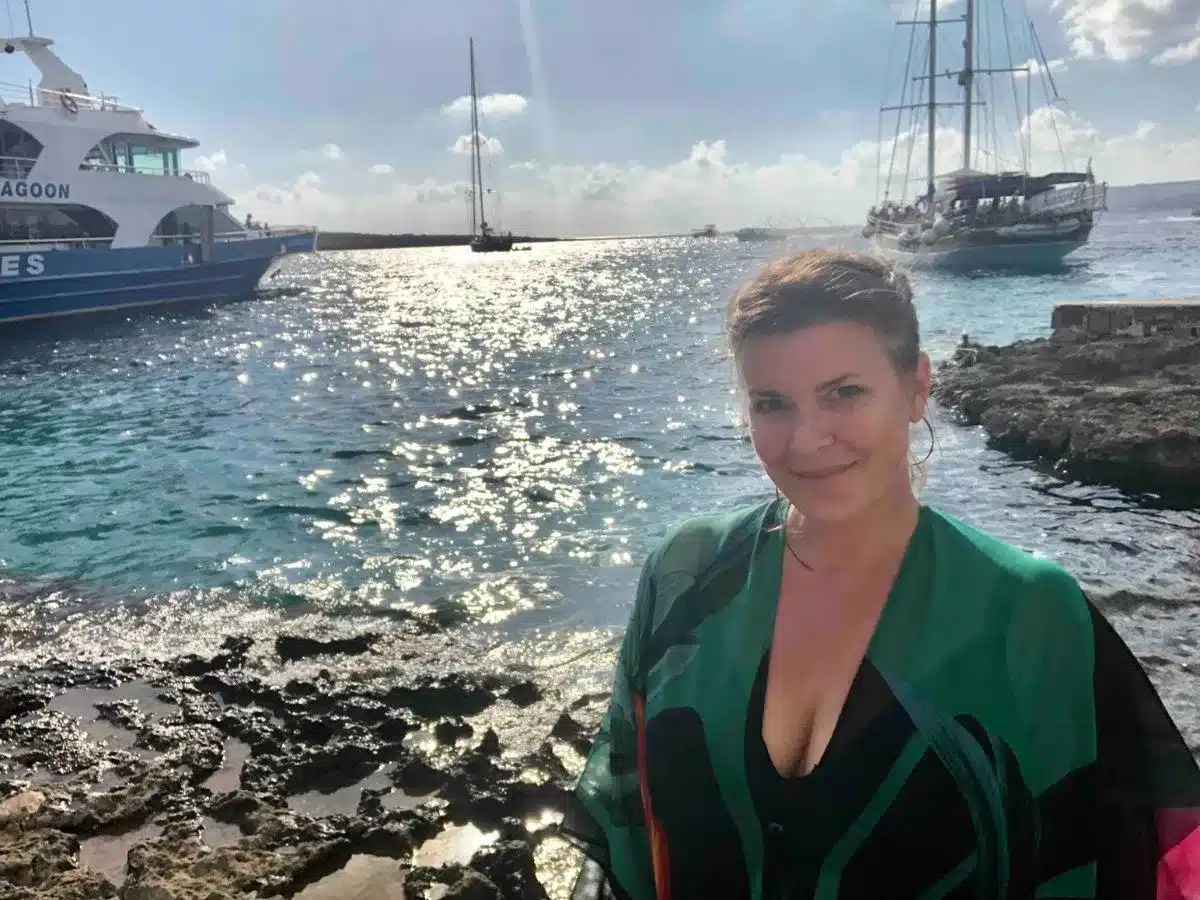 A woman smiles for the camera on a sunny day in Malta, with the glistening Mediterranean Sea behind her. Nearby, a blue and white boat named 'LAGOON' and a classic sailboat add charm to the scenic seascape
